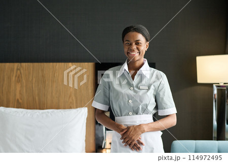 Waist up portrait of young Black woman as housekeeper wearing uniform and smiling at camera standing in hotel room copy space 114972495