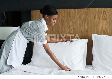 Side view portrait of young African American woman working as housekeeper in hotel and fluffing up fresh pillows copy space 114972497