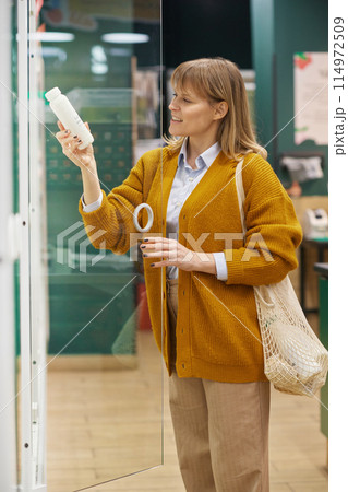 Vertical side view portrait of woman holding milk bottle and smiling while grocery shopping in supermarket Vertical side view portrait of woman holding milk bottle and smiling while grocery shopping in supermarket 114972509