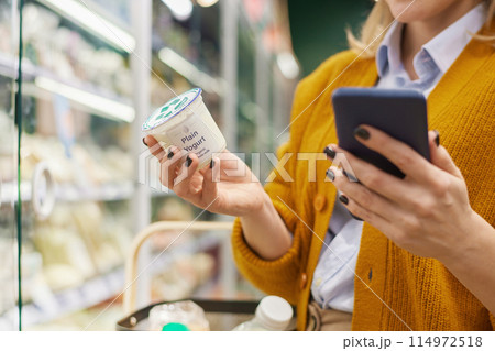Close up of unrecognizable woman buying dairy products in supermarket and using smartphone, copy space 114972518