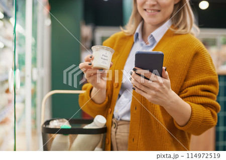 Close up of woman grocery shopping in supermarket and using smartphone in dairy aisle 114972519