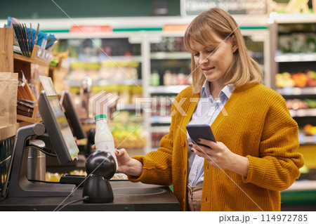 Waist up portrait of blonde adult woman scanning dairy while using self checkout service and holding phone in supermarket 114972785