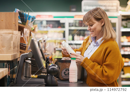 Side view portrait of smiling Caucasian woman using self checkout machine buying milk and groceries in supermarket 114972792