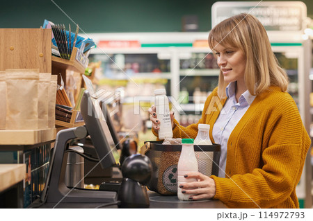 Side view portrait of blonde adult woman using self checkout machine while buying dairy products in supermarket and smiling Side view portrait of blonde adult woman using self checkout machine while buying dairy products in supermarket and smiling 114972793