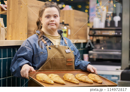 Waist up portrait of confident young woman with Down syndrome working in bakery and looking at camera holding tray with fresh pastry copy space Waist up portrait of confident young woman with Down syndrome working in bakery and looking at camera holding tray with fresh pastry copy space 114972817