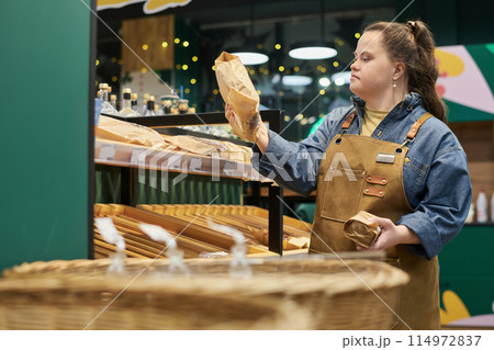 Side view portrait of young woman with Down syndrome stocking shelves in supermarket and holding fresh bread copy space Side view portrait of young woman with Down syndrome stocking shelves in supermarket and holding fresh bread copy space 114972837
