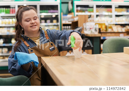 Waist up portrait of smiling young woman with disability working in supermarket and sanitizing tables in cafe area copy space 114972842