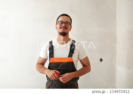 Construction worker in overalls indoor. Smiling young man on white wall background, holding tape measure. cheerful builder with tape measure 114972912