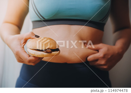 Portrait of obese woman looks sad while pinching her belly fat surrounded by junk foods. Diet fail concept Portrait of obese woman looks sad while pinching her belly fat surrounded by junk foods. Diet fail concept 114974478