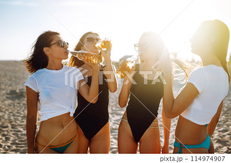 Four beautiful girls cheers and drink beers on the beach, enjoying vacation. Vacation time laughing and smiling. 114974907