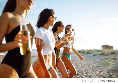 Four beautiful girls cheers and drink beers on the beach, enjoying vacation. Vacation time laughing and smiling. 114974953