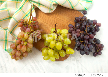 Cutting board with bunch of organic green, pink and blue grapes on white wooden background.. Cutting board with bunch of organic green, pink and blue grapes on white wooden background.. 114975570