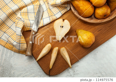Cutting board with whole and slices pears, wicker basket and yellow kitchen towel on white wooden background.. 114975576
