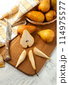 Cutting board with whole and slices pears, wicker basket and yellow kitchen towel on white wooden background.. 114975577