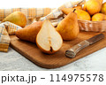 Cutting board with whole and halves pears, wicker basket and yellow kitchen towel on white wooden background.. 114975578