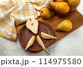 Clay plate with slices and half of pear, wicker basket and yellow kitchen towel on wooden cutting board.. 114975580