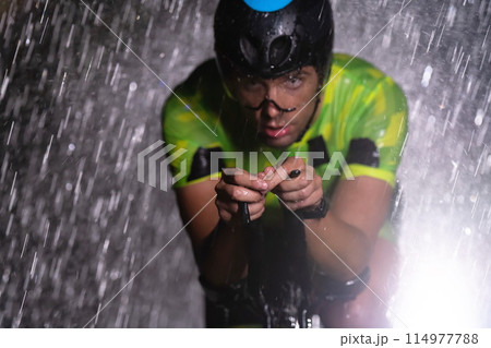 A triathlete braving the rain as he cycles through the night, preparing himself for the upcoming marathon. The blurred raindrops in the foreground and the dark, moody atmosphere in the background add 114977788