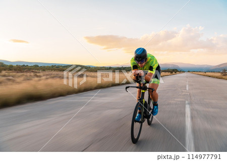 Triathlete riding his bicycle during sunset, preparing for a marathon. The warm colors of the sky provide a beautiful backdrop for his determined and focused effort. 114977791