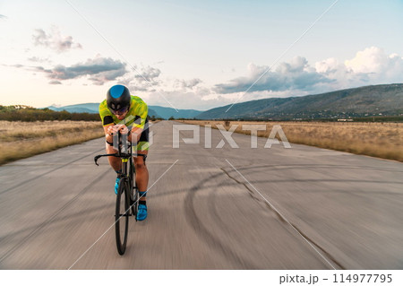 Triathlete riding his bicycle during sunset, preparing for a marathon. The warm colors of the sky provide a beautiful backdrop for his determined and focused effort. 114977795
