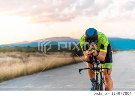 Triathlete riding his bicycle during sunset, preparing for a marathon. The warm colors of the sky provide a beautiful backdrop for his determined and focused effort. 114977804