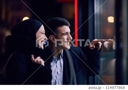 Happy multicultural business couple walking together outdoors in an urban city street at night near a jewelry shopping store window. 114977868
