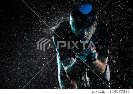A triathlete braving the rain as he cycles through the night, preparing himself for the upcoming marathon. The blurred raindrops in the foreground and the dark, moody atmosphere in the background add A triathlete braving the rain as he cycles through the night, preparing himself for the upcoming marathon. The blurred raindrops in the foreground and the dark, moody atmosphere in the background add 114977885