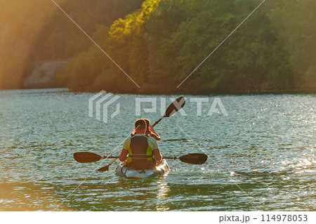 A young couple enjoying an idyllic kayak ride in the middle of a beautiful river surrounded by forest greenery in sunset time  114978053