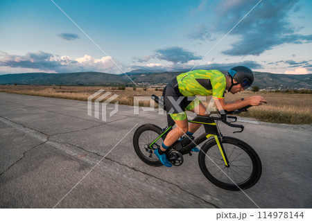 Triathlete riding his bicycle during sunset, preparing for a marathon. The warm colors of the sky provide a beautiful backdrop for his determined and focused effort. 114978144