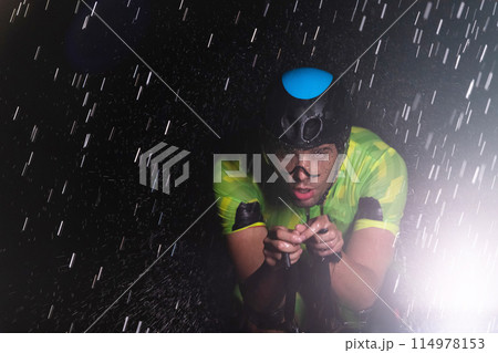 A triathlete braving the rain as he cycles through the night, preparing himself for the upcoming marathon. The blurred raindrops in the foreground and the dark, moody atmosphere in the background add A triathlete braving the rain as he cycles through the night, preparing himself for the upcoming marathon. The blurred raindrops in the foreground and the dark, moody atmosphere in the background add 114978153