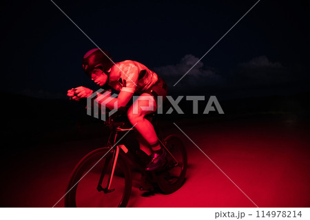 A triathlete rides his bike in the darkness of night, pushing himself to prepare for a marathon. The contrast between the darkness and the light of his bike creates a sense of drama and highlights the 114978214