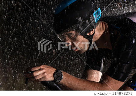 A triathlete braving the rain as he cycles through the night, preparing himself for the upcoming marathon. The blurred raindrops in the foreground and the dark, moody atmosphere in the background add 114978273