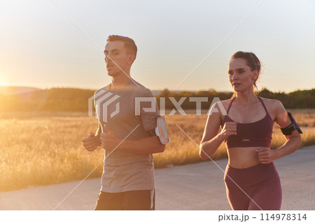 A handsome young couple running together during the early morning hours, with the mesmerizing sunrise casting a warm glow, symbolizing their shared love and vitality 114978314