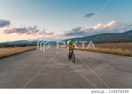 Triathlete riding his bicycle during sunset, preparing for a marathon. The warm colors of the sky provide a beautiful backdrop for his determined and focused effort. 114978399