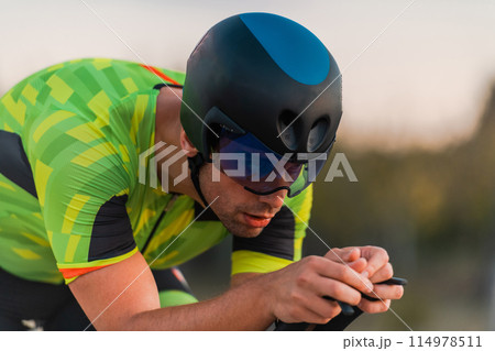 Close up photo of triathlete riding his bicycle during sunset, preparing for a marathon. The warm colors of the sky provide a beautiful backdrop for his determined and focused effort. 114978511