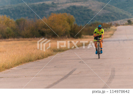 Triathlete riding his bicycle during sunset, preparing for a marathon. The warm colors of the sky provide a beautiful backdrop for his determined and focused effort. Triathlete riding his bicycle during sunset, preparing for a marathon. The warm colors of the sky provide a beautiful backdrop for his determined and focused effort. 114978642