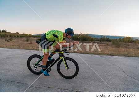 Triathlete riding his bicycle during sunset, preparing for a marathon. The warm colors of the sky provide a beautiful backdrop for his determined and focused effort. Triathlete riding his bicycle during sunset, preparing for a marathon. The warm colors of the sky provide a beautiful backdrop for his determined and focused effort. 114978677