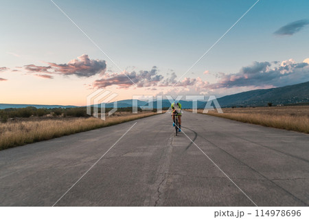 Triathlete riding his bicycle during sunset, preparing for a marathon. The warm colors of the sky provide a beautiful backdrop for his determined and focused effort. Triathlete riding his bicycle during sunset, preparing for a marathon. The warm colors of the sky provide a beautiful backdrop for his determined and focused effort. 114978696