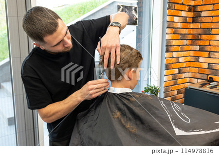 Young male barber making a haircut for cute little boy in the modern barbershop. Child in barbershop. Haircuts for Kids 114978816