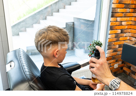 A boy sits in a barbershop while he is sprayed with water to start a haircut. A master in a barbershop sprays water on a boy who is sitting and waiting for his haircut.  114978824