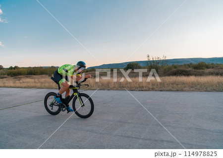 Triathlete riding his bicycle during sunset, preparing for a marathon. The warm colors of the sky provide a beautiful backdrop for his determined and focused effort. 114978825