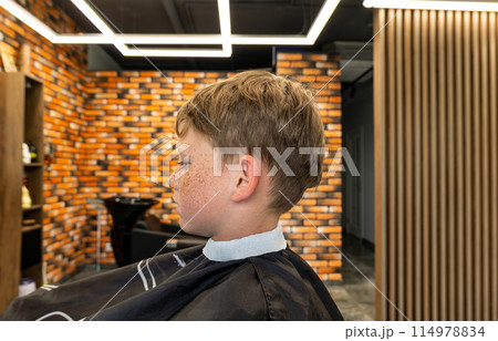 The picture of a small child before being cut was taken in a barbshop studio, the child has very beautiful blond hair that turns golden. The child is happy that he will have a new style of cutting. 114978834