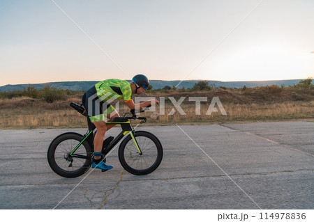 Triathlete riding his bicycle during sunset, preparing for a marathon. The warm colors of the sky provide a beautiful backdrop for his determined and focused effort. 114978836