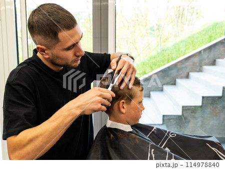 Man barber cutting little boy's hair using comb and scissors. Child getting haircut from adult male, likely barber. Professional hairdresser and cute client at modern barbershop. Man barber cutting little boy's hair using comb and scissors. Child getting haircut from adult male, likely barber. Professional hairdresser and cute client at modern barbershop. 114978840