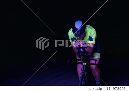 A triathlete rides his bike in the darkness of night, pushing himself to prepare for a marathon. The contrast between the darkness and the light of his bike creates a sense of drama and highlights the A triathlete rides his bike in the darkness of night, pushing himself to prepare for a marathon. The contrast between the darkness and the light of his bike creates a sense of drama and highlights the 114978903