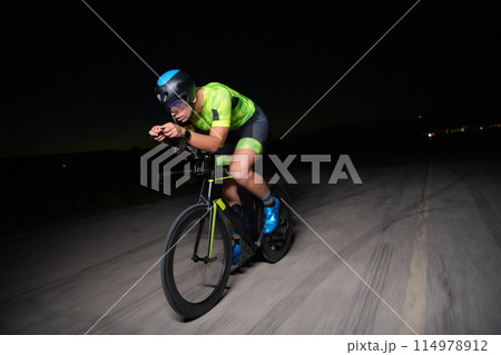 A triathlete rides his bike in the darkness of night, pushing himself to prepare for a marathon. The contrast between the darkness and the light of his bike creates a sense of drama and highlights the 114978912