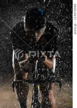 A triathlete braving the rain as he cycles through the night, preparing himself for the upcoming marathon. The blurred raindrops in the foreground and the dark, moody atmosphere in the background add 114978980