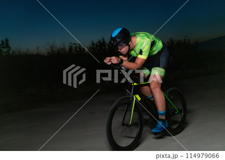 A triathlete rides his bike in the darkness of night, pushing himself to prepare for a marathon. The contrast between the darkness and the light of his bike creates a sense of drama and highlights the A triathlete rides his bike in the darkness of night, pushing himself to prepare for a marathon. The contrast between the darkness and the light of his bike creates a sense of drama and highlights the 114979066