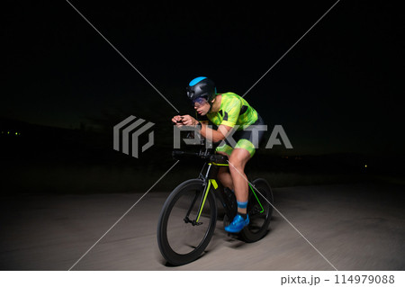 A triathlete rides his bike in the darkness of night, pushing himself to prepare for a marathon. The contrast between the darkness and the light of his bike creates a sense of drama and highlights the A triathlete rides his bike in the darkness of night, pushing himself to prepare for a marathon. The contrast between the darkness and the light of his bike creates a sense of drama and highlights the 114979088
