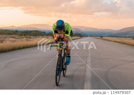 Triathlete riding his bicycle during sunset, preparing for a marathon. The warm colors of the sky provide a beautiful backdrop for his determined and focused effort. Triathlete riding his bicycle during sunset, preparing for a marathon. The warm colors of the sky provide a beautiful backdrop for his determined and focused effort. 114979307