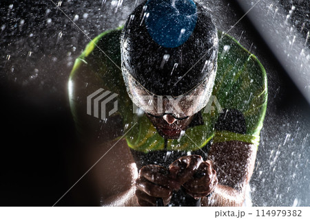 A triathlete braving the rain as he cycles through the night, preparing himself for the upcoming marathon. The blurred raindrops in the foreground and the dark, moody atmosphere in the background add A triathlete braving the rain as he cycles through the night, preparing himself for the upcoming marathon. The blurred raindrops in the foreground and the dark, moody atmosphere in the background add 114979382